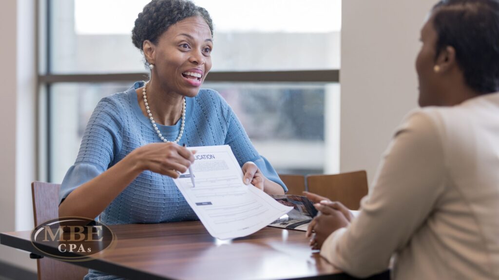 A woman in a blue top reviewing application documents with a client at a wooden desk with MBE CPAs logo.