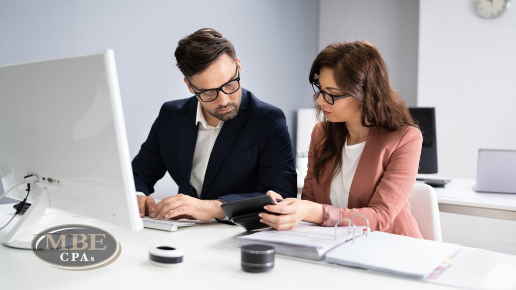 Two business professionals in glasses reviewing documents together at a desk with a computer and MBE CPAs logo.