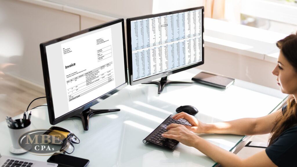 A woman working at a glass desk using dual monitors to view an invoice and spreadsheet, with MBE CPAs logo.