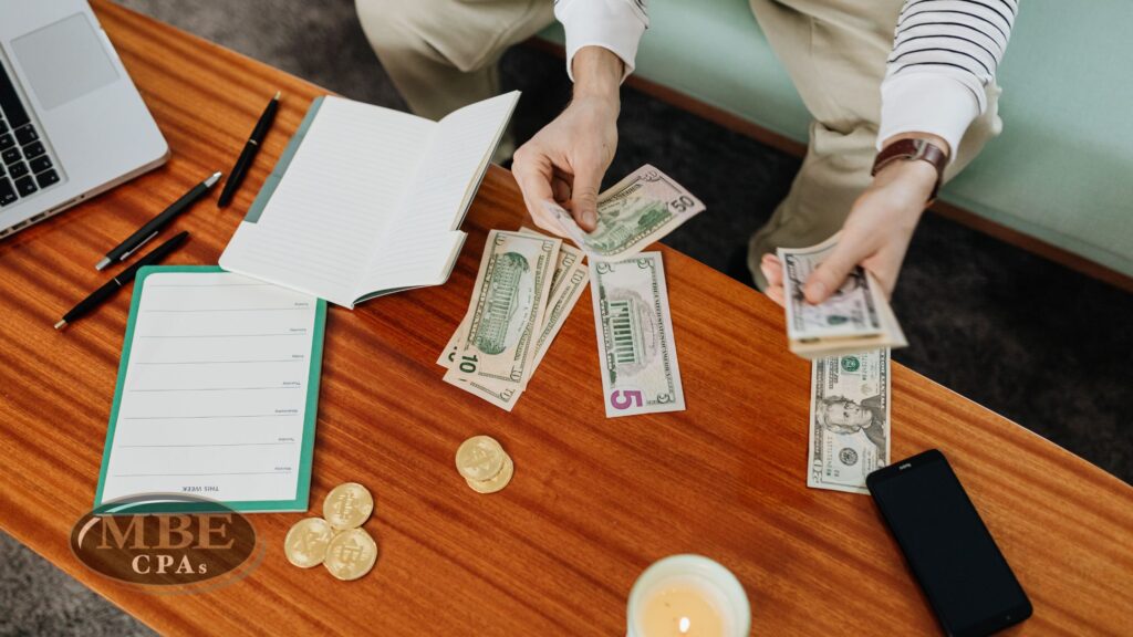 A person counting cash on a wooden desk near notebooks, gold coins, a laptop, and the MBE CPAs logo.