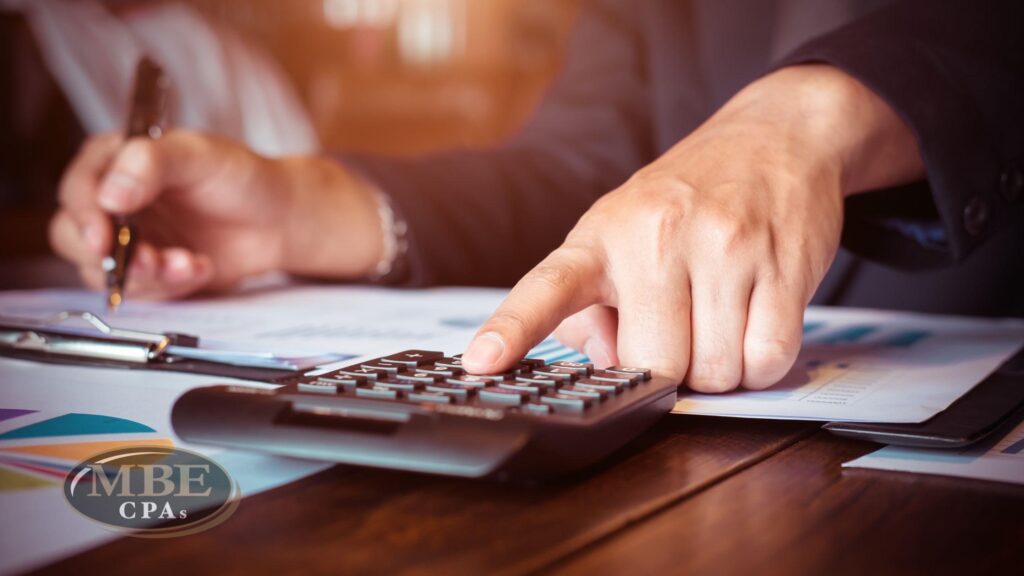 Close-up of a person using a calculator and pen to analyze financial documents with the MBE CPAs logo.