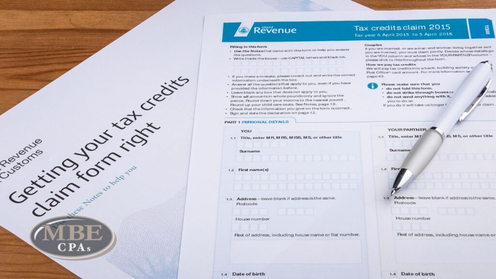 Tax credit claim paperwork and a silver pen resting on a wooden desk, featuring the MBE CPAs logo overlay.