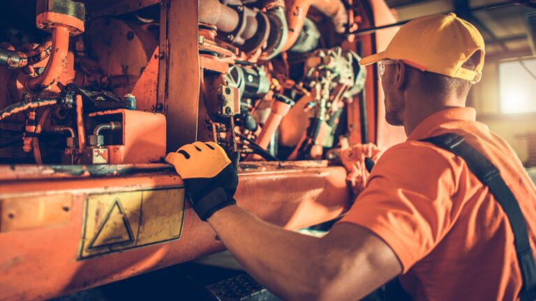 A technician wearing a yellow cap and work gloves inspecting large orange heavy machinery in a workshop.