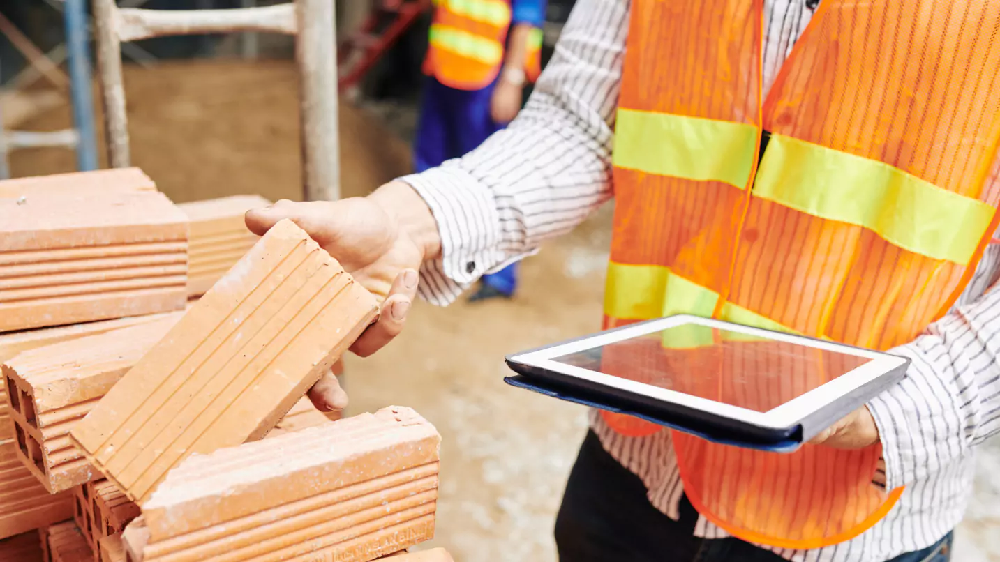 A construction worker holding a tablet and bricks while using construction equipment.