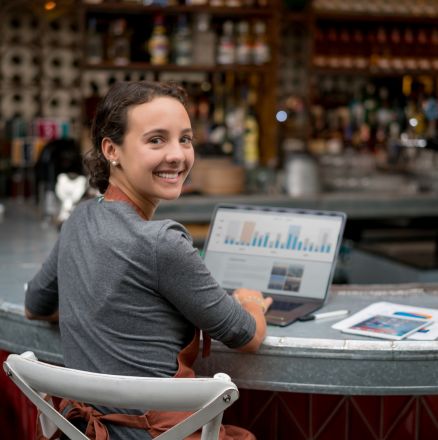 A smiling restaurant manager using a laptop displaying financial charts while seated at a bar counter.