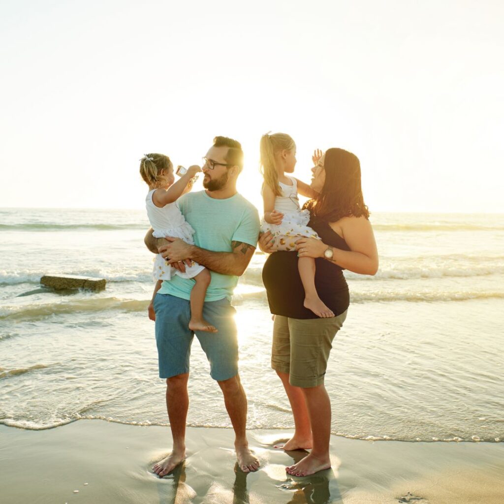 A pregnant mother and father holding their two young daughters while standing on a sandy beach at sunset.