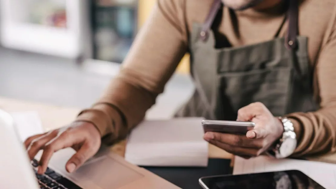 A man is holding a phone while typing on a laptop