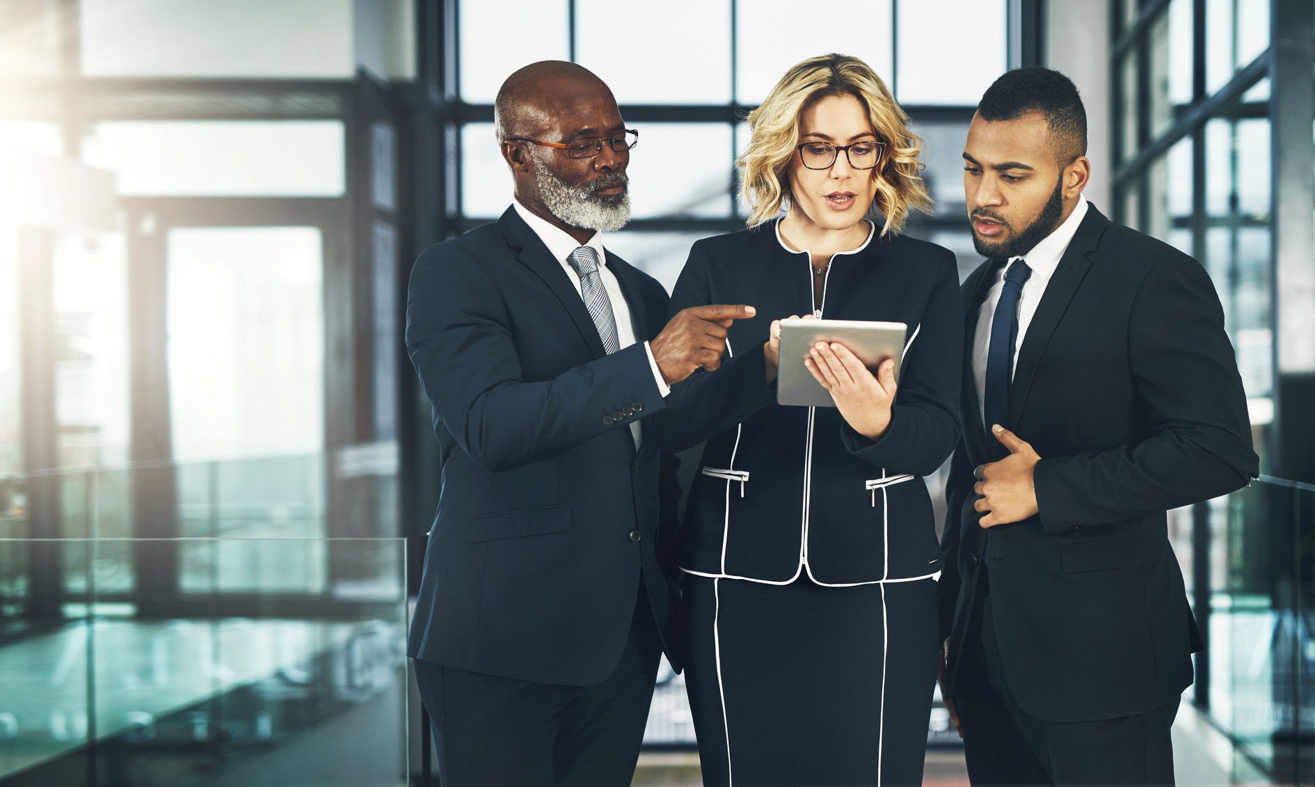 Three business professionals in suits analyzing digital information on a tablet in a modern office hall.