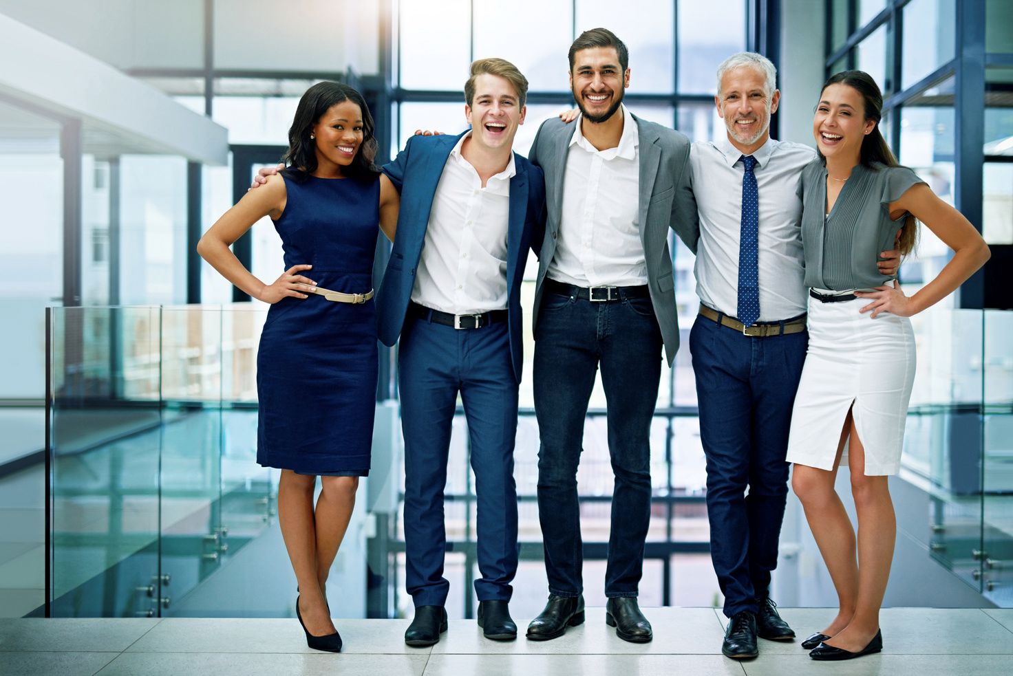 A smiling team of five colleagues posing together full-length inside a modern glass office building.