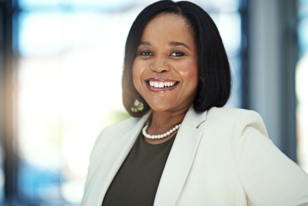 A professional headshot of a smiling woman in a white blazer and pearl necklace in a brightly lit office.