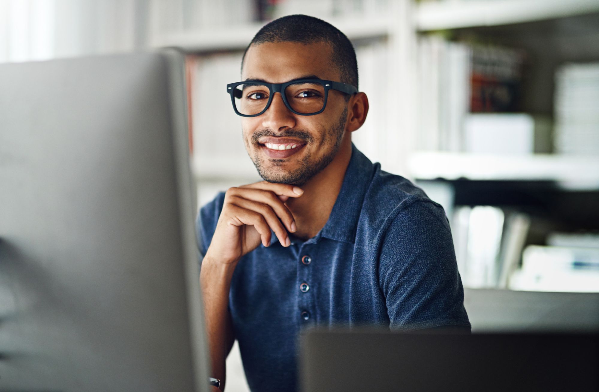 A man in dark-rimmed glasses looking at the camera with a smile while sitting at a workstation with a monitor.