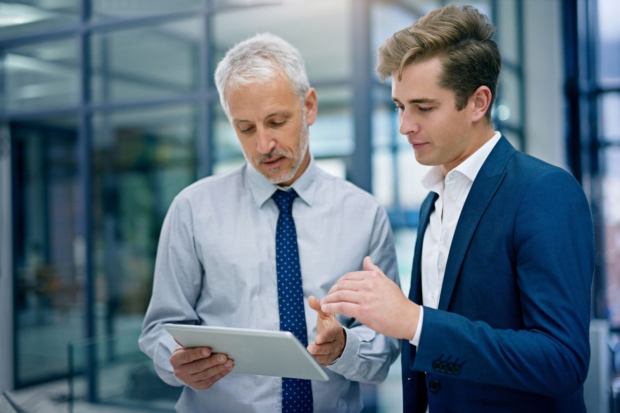 Two businessmen standing together in a modern office, analyzing information on a tablet computer together.