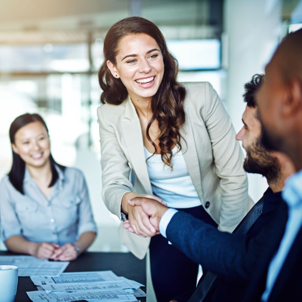 A smiling businesswoman in a blazer shaking hands with a colleague across a conference table during a meeting.