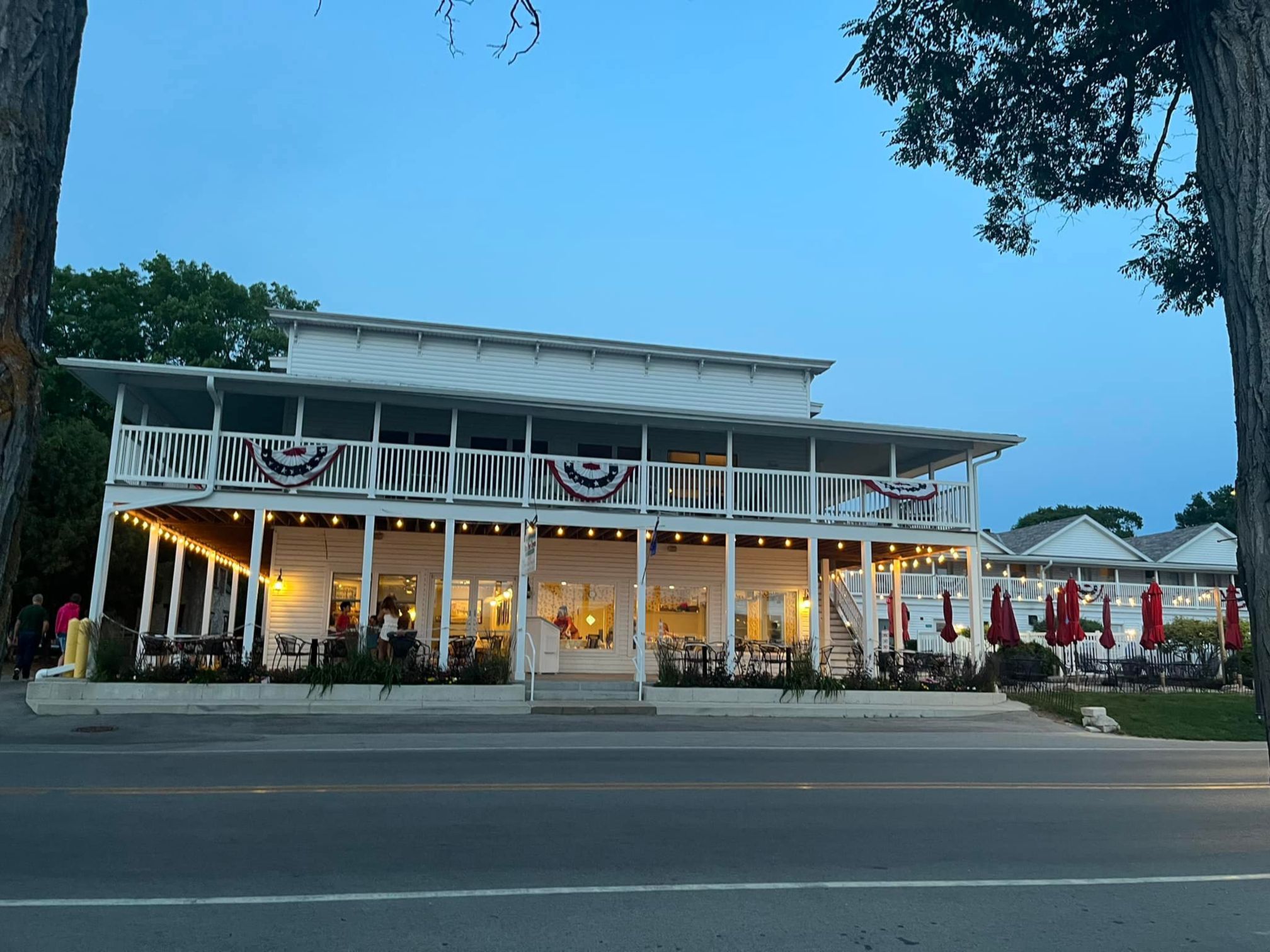 A white two-story historic inn and restaurant decorated with patriotic bunting and string lights at dusk.