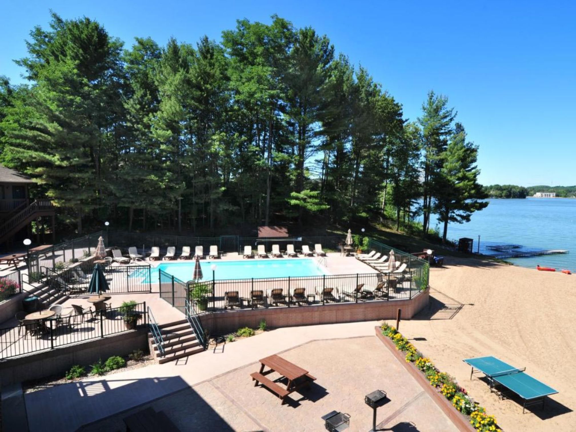 An outdoor resort swimming pool and sun deck beside a sandy beach and lake, surrounded by pine trees.