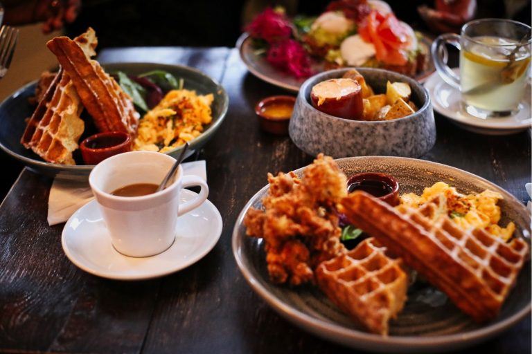 Table spread with plates of Belgian waffles, fried chicken, scrambled eggs, roasted potatoes, and coffee.