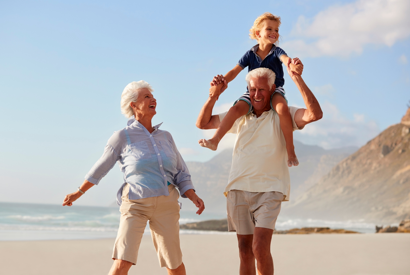 An elderly man and woman walking on a beach. The man is carrying his grandchild on his shoulders.