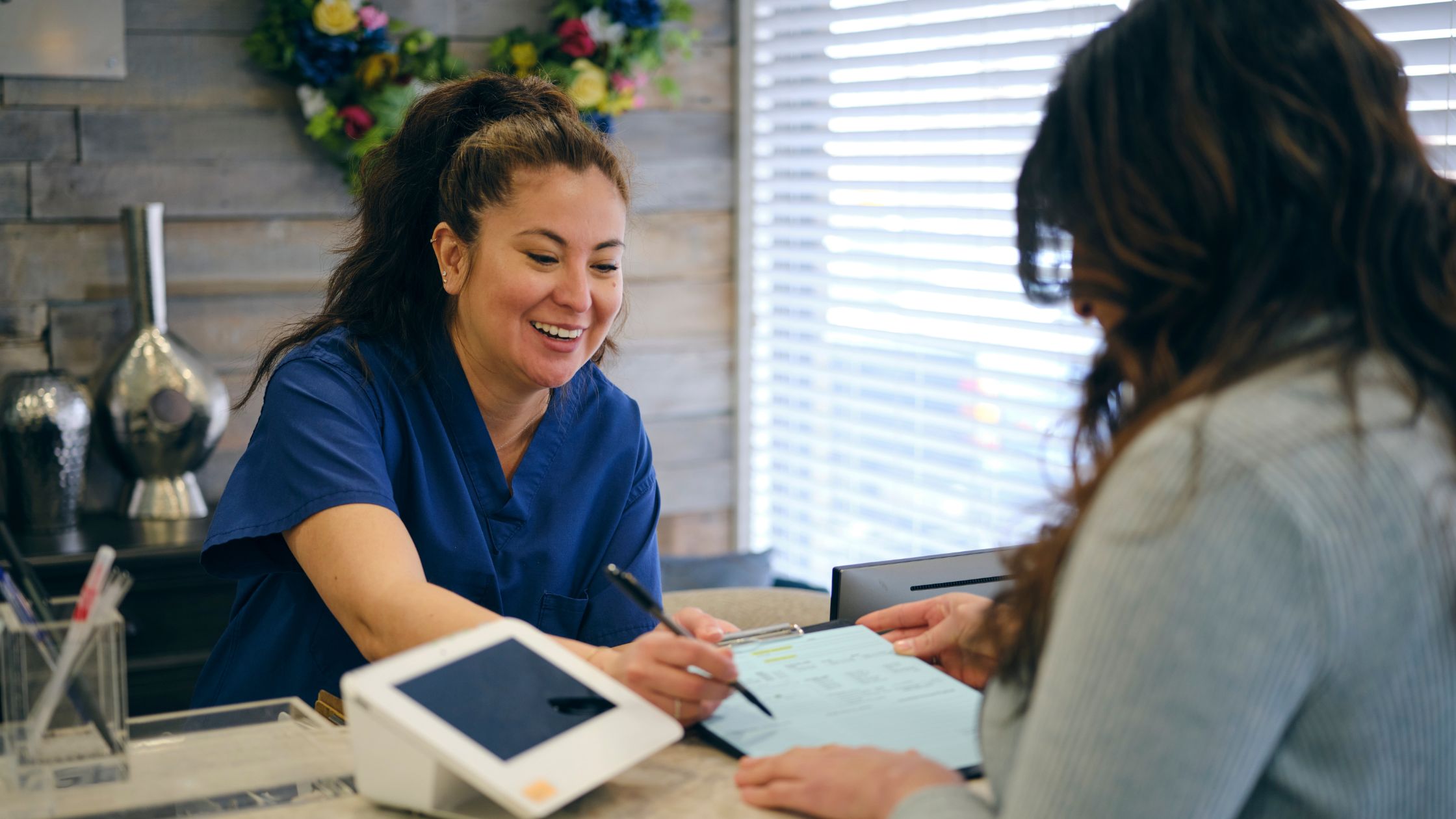 A healthcare professional in blue scrubs reviews paperwork with a patient at a reception desk
